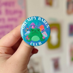 May include: A blue and green button with a frog surrounded by mushrooms and the text "Grumpy Babes Club".