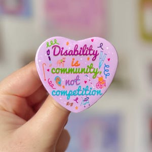 May include: A pink heart-shaped button with the text "Disability is community, not competition" in blue and purple lettering. The button is decorated with colorful illustrations of accessibility aids, including wheelchairs, crutches, and hearing aids.