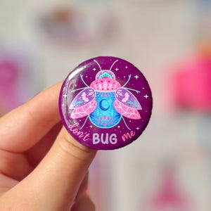 May include: A round, purple button with a whimsical bug illustration. The bug features pink, blue, and teal details, with a crescent moon design. The text "don't BUG me" is written in white. The button has a glossy finish.
