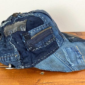 May include: A baseball cap constructed from various shades of blue denim fabric patches. The cap features visible white decorative stitching, a dark blue zipper detail on the side, and a rectangular grey fabric patch with the text 781-5A printed on it.