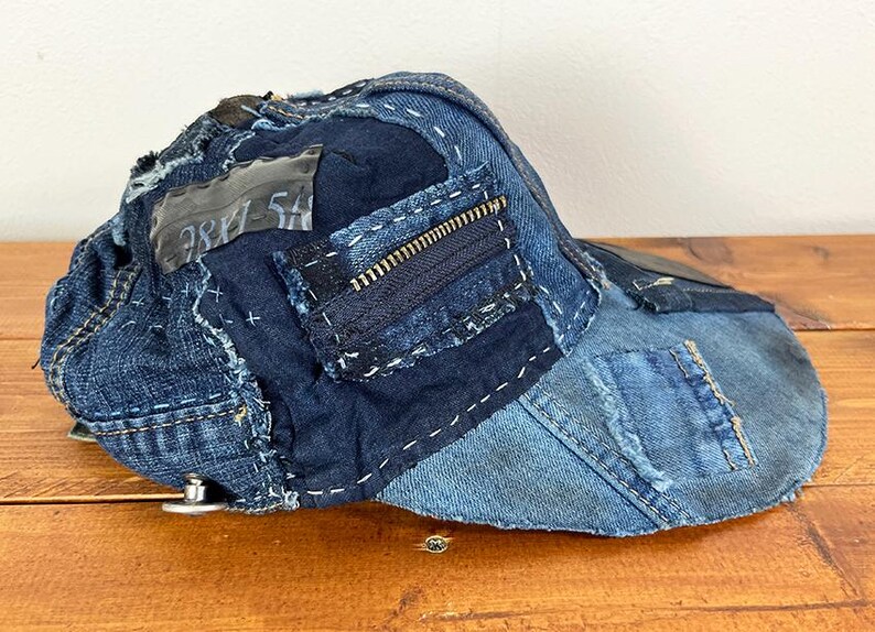 May include: A baseball cap constructed from various shades of blue denim fabric patches. The cap features visible white decorative stitching, a dark blue zipper detail on the side, and a rectangular grey fabric patch with the text 781-5A printed on it.