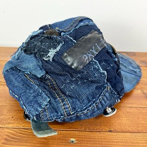 May include: A baseball cap constructed from various shades of blue denim fabric patches. The cap features visible white sashiko-style stitching, a dark rectangular patch with the text 28X1, and metallic silver buttons along the lower edge.