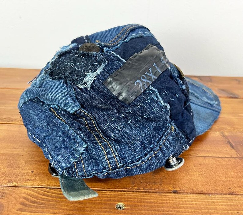 May include: A baseball cap constructed from various shades of blue denim fabric patches. The cap features visible white sashiko-style stitching, a dark rectangular patch with the text 28X1, and metallic silver buttons along the lower edge.