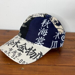 May include: A baseball cap featuring a patchwork design of cream, black, and navy blue fabric panels. The cap is decorated with black Japanese kanji characters and text, including the numbers 92-00, printed across the front and side panels.