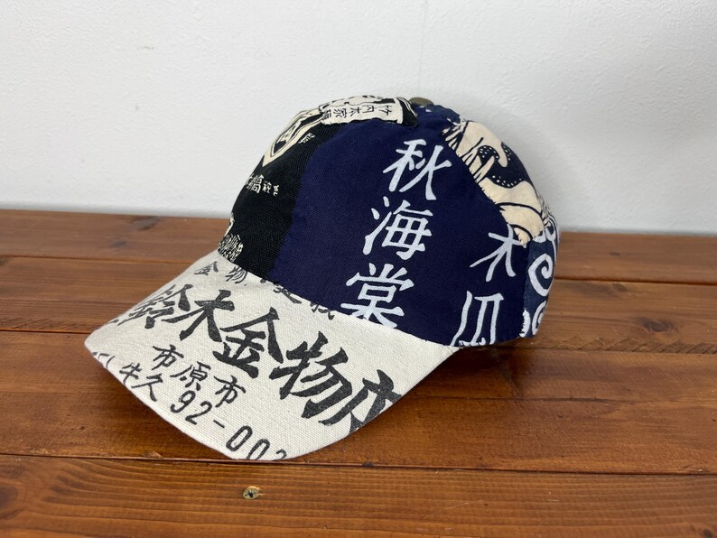 May include: A baseball cap featuring a patchwork design of cream, black, and navy blue fabric panels. The cap is decorated with black Japanese kanji characters and text, including the numbers 92-00, printed across the front and side panels.