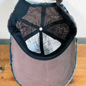 May include: A blue denim baseball cap with a brown fabric lining and a white button on the top. The cap has a distressed look with frayed edges.