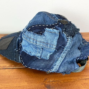 May include: A baseball cap constructed from various shades of blue denim fabric. The cap features a patchwork design with visible white stitching, frayed edges, and multiple denim panels layered together to form the crown and the brim.