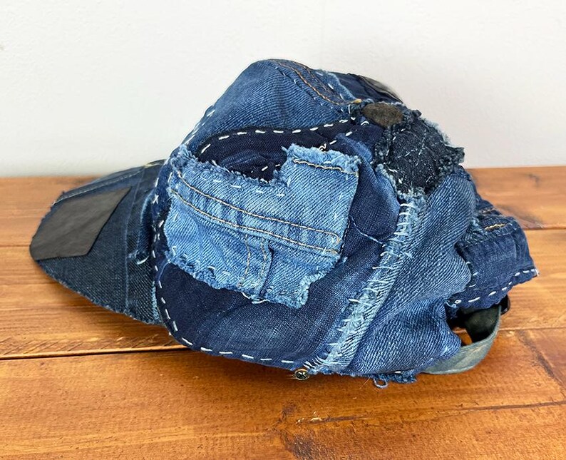 May include: A baseball cap constructed from various shades of blue denim fabric. The cap features a patchwork design with visible white stitching, frayed edges, and multiple denim panels layered together to form the crown and the brim.