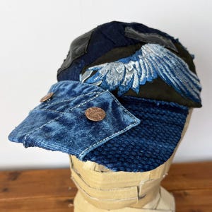 Op de afbeelding: Een blauwe denim en patchwork baseballcap met een geborduurd vleugelontwerp. De pet heeft een verweerde look en heeft een koperen knoop met de tekst "Off the Grid" aan de voorkant.