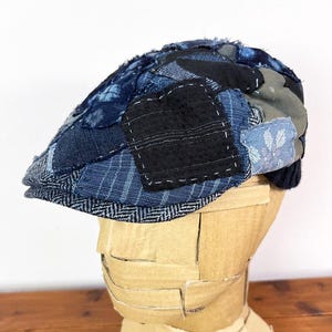 May include: A blue and grey patchwork newsboy cap made from different fabrics. The cap has a unique and textured design.