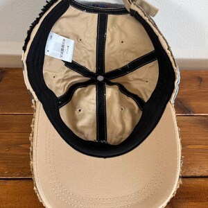 Op de afbeelding: Een beige baseballpet met een zwarte klep en een rafelige rand. De binnenkant van de pet is beige met zwarte stiksels en een zwarte band. De pet heeft een label met de tekst "サイズ調整可能".