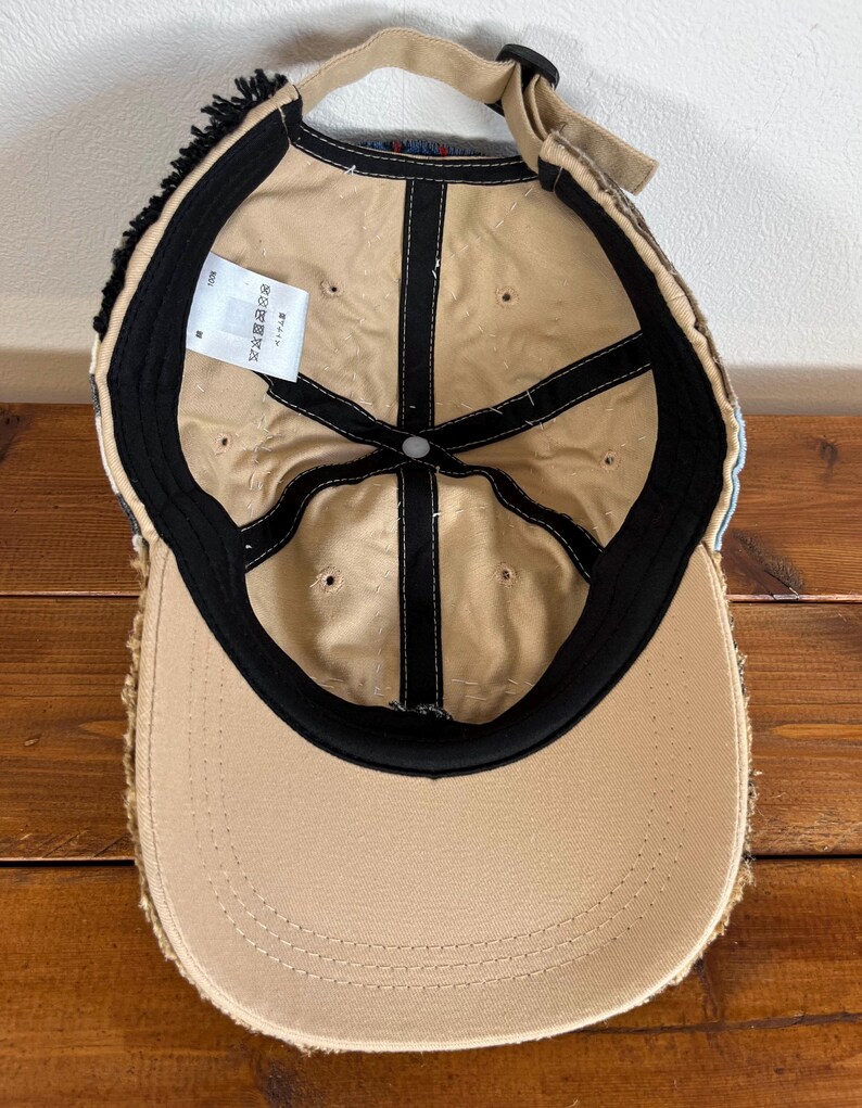 Op de afbeelding: Een beige baseballpet met een zwarte klep en een rafelige rand. De binnenkant van de pet is beige met zwarte stiksels en een zwarte band. De pet heeft een label met de tekst "サイズ調整可能".