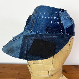May include: A baseball cap constructed from various shades of blue denim fabric patches. The cap features visible white cross-stitch embroidery on the crown, frayed edges, and a solid black rectangular patch on the front brim section.