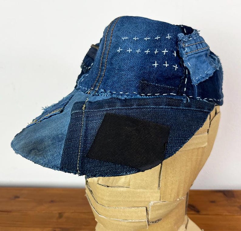 May include: A baseball cap constructed from various shades of blue denim fabric patches. The cap features visible white cross-stitch embroidery on the crown, frayed edges, and a solid black rectangular patch on the front brim section.