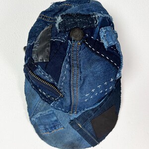 May include: A baseball cap constructed from various shades of blue denim fabric, featuring visible frayed edges, white decorative stitching, a dark metal zipper detail, a black rectangular patch, and a central dark button on the crown.
