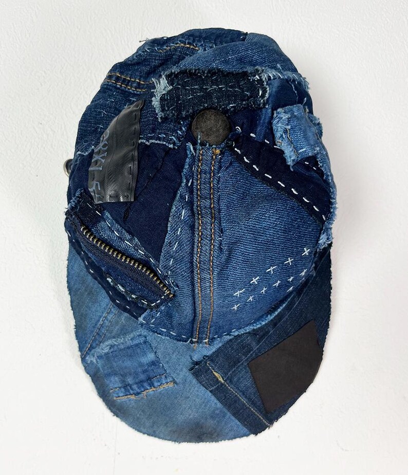May include: A baseball cap constructed from various shades of blue denim fabric, featuring visible frayed edges, white decorative stitching, a dark metal zipper detail, a black rectangular patch, and a central dark button on the crown.
