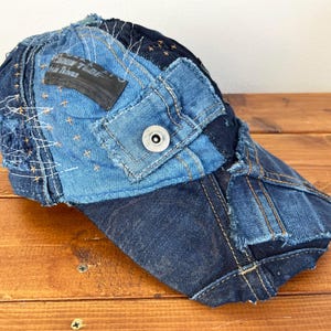 Op de afbeelding: Een blauwe denim baseballcap met een patchwork ontwerp. De cap is gemaakt van verschillende stukken denim die aan elkaar zijn genaaid, wat een unieke en stijlvolle look cre&euml;ert. De cap heeft een zwarte klep en een kleine metalen knop bovenop.