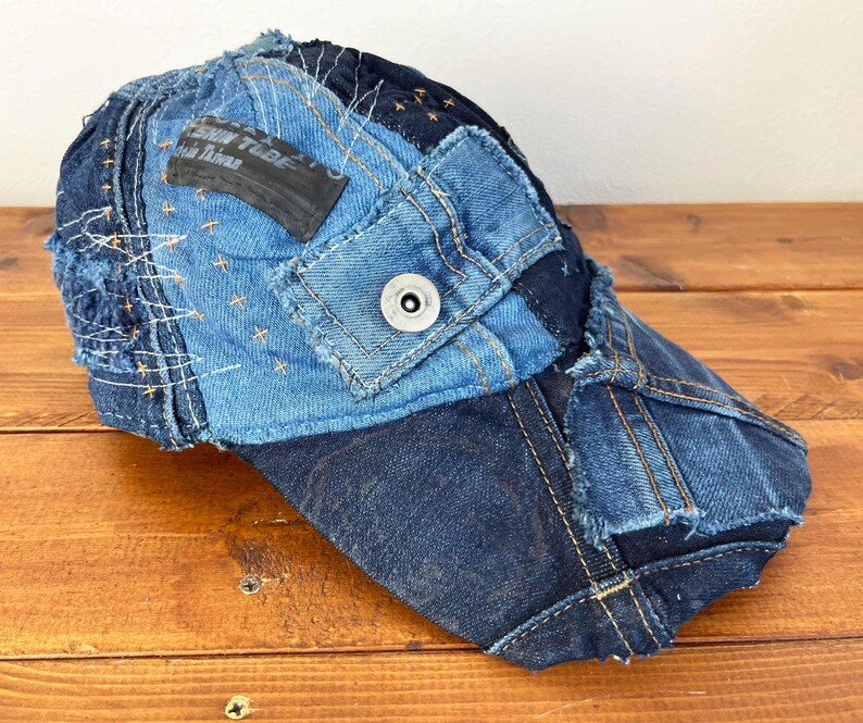 Op de afbeelding: Een blauwe denim baseballcap met een patchwork ontwerp. De cap is gemaakt van verschillende stukken denim die aan elkaar zijn genaaid, wat een unieke en stijlvolle look cre&euml;ert. De cap heeft een zwarte klep en een kleine metalen knop bovenop.