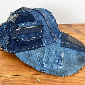 May include: A baseball cap constructed from various shades of blue denim fabric. The cap features a patchwork design with visible white stitching, frayed edges, a dark blue zipper detail on the side, and a smooth dark patch on the curved brim.