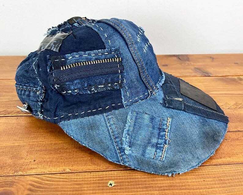 May include: A baseball cap constructed from various shades of blue denim fabric. The cap features a patchwork design with visible white stitching, frayed edges, a dark blue zipper detail on the side, and a smooth dark patch on the curved brim.
