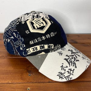 May include: A baseball cap featuring a patchwork design with navy blue, black, and cream panels. The cap displays Japanese kanji characters and graphic patterns, including a prominent central emblem and text on the cream-coloured curved brim.