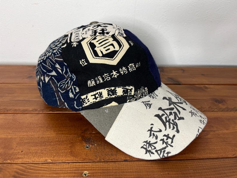 May include: A baseball cap featuring a patchwork design with navy blue, black, and cream panels. The cap displays Japanese kanji characters and graphic patterns, including a prominent central emblem and text on the cream-coloured curved brim.