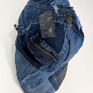 May include: A baseball cap constructed from repurposed blue denim fabric featuring visible white sashiko-style stitching, frayed edges, a dark zipper detail, and a rectangular black patch with the text 38X154 printed in white.
