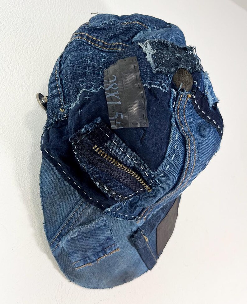 May include: A baseball cap constructed from repurposed blue denim fabric featuring visible white sashiko-style stitching, frayed edges, a dark zipper detail, and a rectangular black patch with the text 38X154 printed in white.
