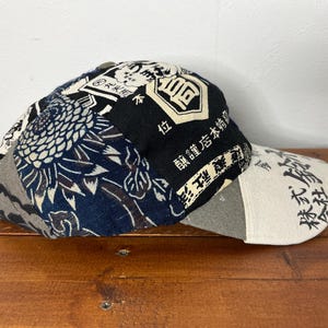 May include: A baseball cap featuring a patchwork design of dark blue and white fabric with floral patterns and black panels printed with Japanese kanji characters. The brim is a light cream colour with black Japanese text printed across the surface.