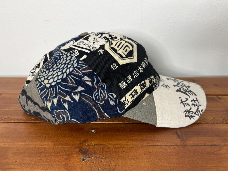 May include: A baseball cap featuring a patchwork design of dark blue and white fabric with floral patterns and black panels printed with Japanese kanji characters. The brim is a light cream colour with black Japanese text printed across the surface.
