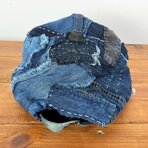 May include: A baseball cap constructed from various shades of blue denim fabric patches. The cap features visible frayed edges, decorative white cross-stitch embroidery, and a dark grey leather-like patch on the side with a button at the top.