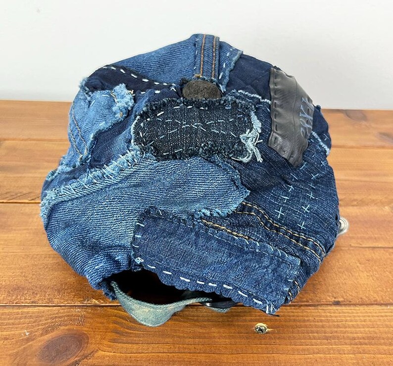 May include: A baseball cap constructed from various shades of blue denim fabric patches. The cap features visible frayed edges, decorative white cross-stitch embroidery, and a dark grey leather-like patch on the side with a button at the top.