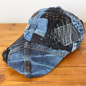 Op de afbeelding: Een blauwe denim baseballcap met een patchwork ontwerp. De cap is gemaakt van verschillende stukken denim die aan elkaar zijn genaaid, en de naden zijn zichtbaar. De cap heeft een rafelige rand en een knoop aan de voorkant.