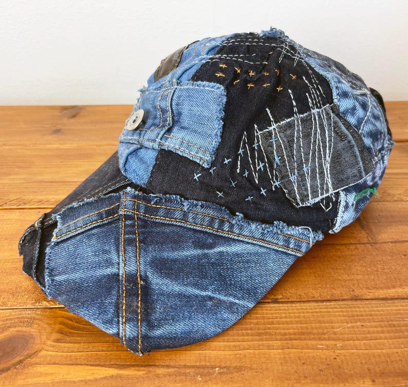 Op de afbeelding: Een blauwe denim baseballcap met een patchwork ontwerp. De cap is gemaakt van verschillende stukken denim die aan elkaar zijn genaaid, en de naden zijn zichtbaar. De cap heeft een rafelige rand en een knoop aan de voorkant.