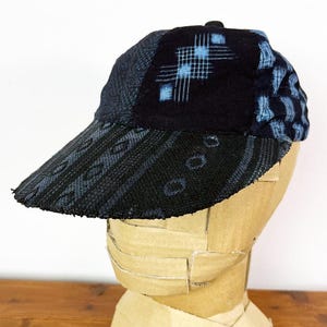 May include: A baseball cap constructed from various panels of dark blue and black fabric. The front panel features a geometric grid pattern in light blue, while the brim and side panels display textured, woven patterns in shades of black and blue.