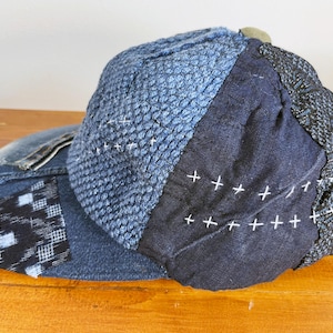 May include: A blue denim baseball cap with a patchwork design. The cap is made of different shades of blue denim and has white stitching. The cap has a black and white checkered patch on the side.