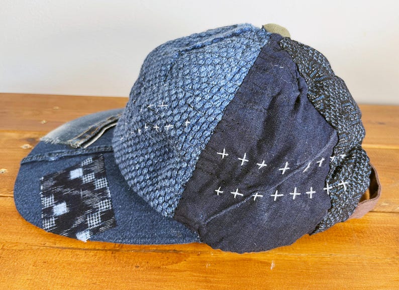 May include: A blue denim baseball cap with a patchwork design. The cap is made of different shades of blue denim and has white stitching. The cap has a black and white checkered patch on the side.