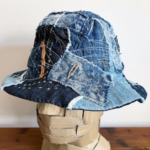 May include: A blue denim bucket hat made from patchwork fabric with visible stitching. The hat is made from different shades of blue denim and has a wide brim.