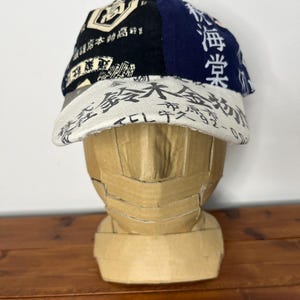 May include: A baseball cap featuring a multi-panel design with black, navy blue, and off-white fabric sections. The cap is covered in various Japanese kanji characters and text prints in black and white, including a prominent brim with text.