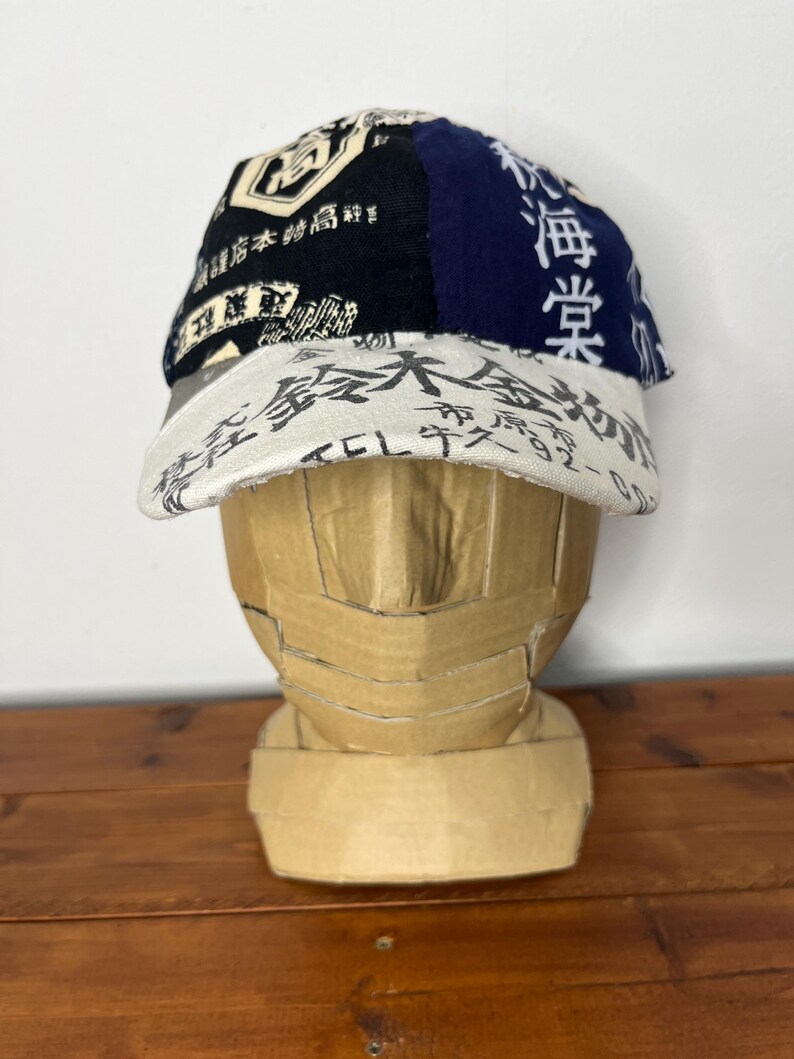 May include: A baseball cap featuring a multi-panel design with black, navy blue, and off-white fabric sections. The cap is covered in various Japanese kanji characters and text prints in black and white, including a prominent brim with text.