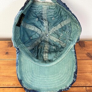Op de afbeelding: Een blauwe denim baseballpet met een verweerde look. De pet heeft een gescheurde rand en een verbleekte blauwe kleur. De binnenkant van de pet is gevoerd met een lichtblauwe stof.