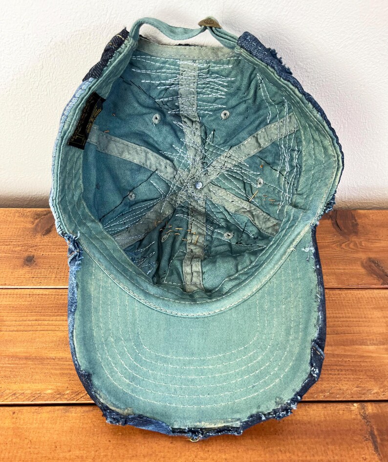 Op de afbeelding: Een blauwe denim baseballpet met een verweerde look. De pet heeft een gescheurde rand en een verbleekte blauwe kleur. De binnenkant van de pet is gevoerd met een lichtblauwe stof.