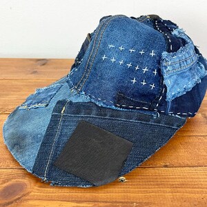May include: A baseball cap constructed from various shades of blue denim fabric patches. The cap features visible white sashiko-style stitching in a cross pattern, frayed edges, and a solid black square patch on the side of the brim.