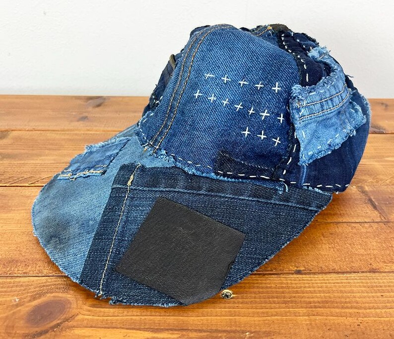 May include: A baseball cap constructed from various shades of blue denim fabric patches. The cap features visible white sashiko-style stitching in a cross pattern, frayed edges, and a solid black square patch on the side of the brim.
