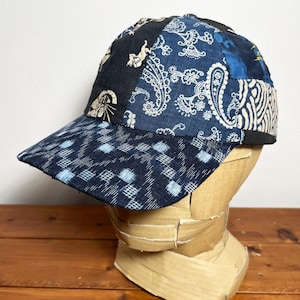 May include: A baseball cap constructed from a patchwork of various blue and black fabrics. The crown features panels with paisley, geometric, and animal motifs, while the brim displays a dark blue fabric with a light blue checkered pattern.