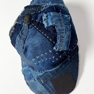 May include: A baseball cap constructed from various shades of blue denim fabric. The cap features visible frayed edges, decorative white cross-stitch embroidery, and a black rectangular patch on the brim. The design includes multiple denim panels.
