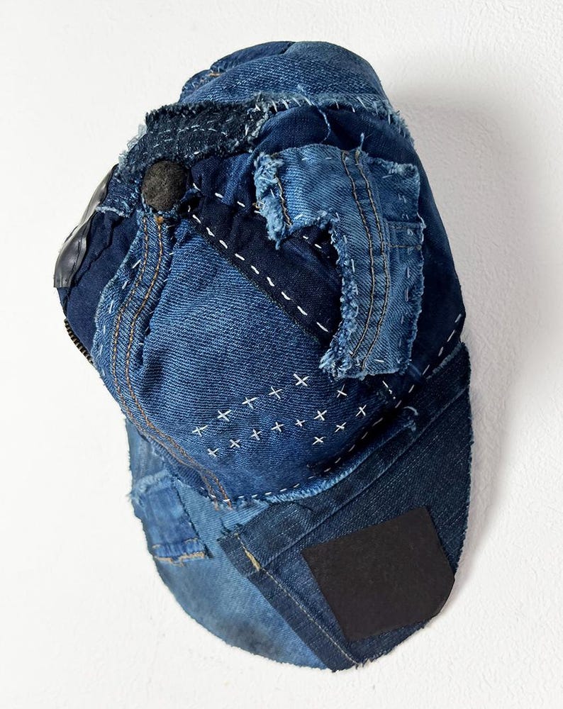 May include: A baseball cap constructed from various shades of blue denim fabric. The cap features visible frayed edges, decorative white cross-stitch embroidery, and a black rectangular patch on the brim. The design includes multiple denim panels.