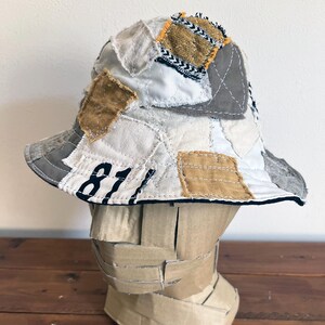 May include: A white bucket hat with a patchwork design featuring various shades of brown and gray fabric. The hat has a black brim and the number "81" is stitched on the front.