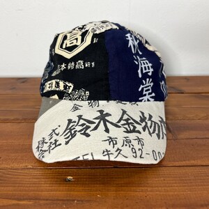 May include: A baseball cap featuring a patchwork design with black, navy blue, and off-white panels. The cap is covered in various Japanese kanji characters and text, including the prominent black text on the off-white brim and crown panels.
