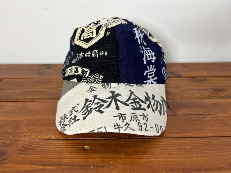 May include: A baseball cap featuring a patchwork design with black, navy blue, and off-white panels. The cap is covered in various Japanese kanji characters and text, including the prominent black text on the off-white brim and crown panels.